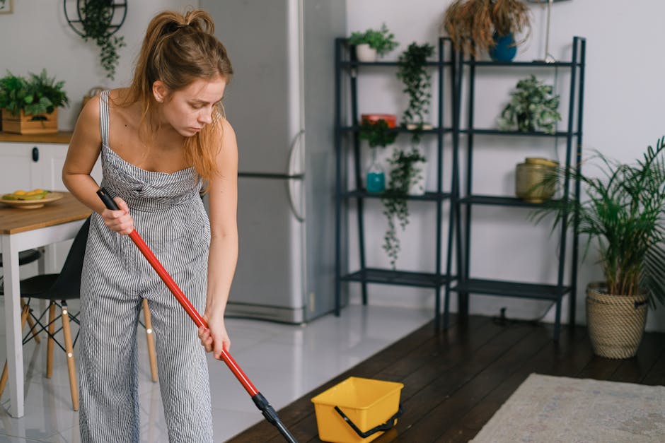 A young woman in a jumpsuit mopping a modern kitchen floor with a red mop, surrounded by plants.